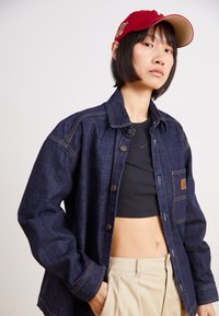 Chaqueta de denim en azul oscuro con costuras amarillas en contraste, presenta un cierre de botones en la parte delantera y un bolsillo en el pecho. Llevada sobre un top corto negro.