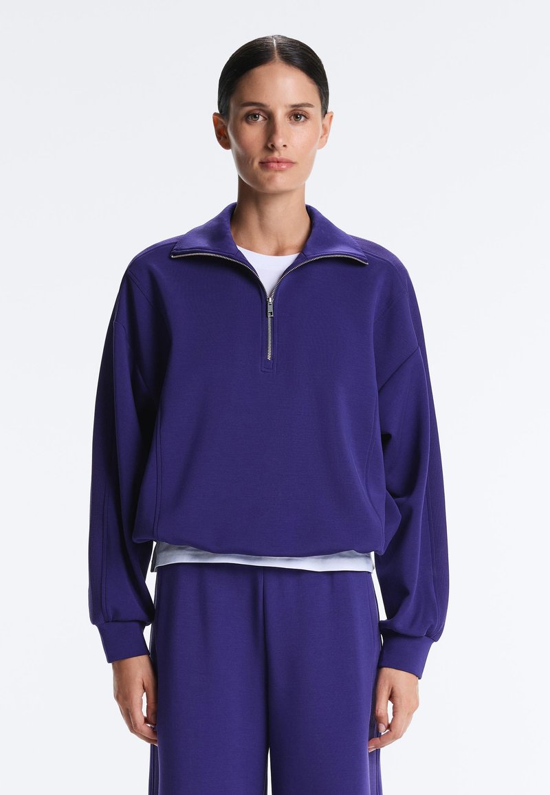 Kvinde med mørkt hår iført en løs lilla sweatshirt med lynlås og matchende bukser, stående mod en ensfarvet hvid baggrund.