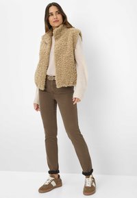 Beige nepleren vest, lange mouwen crèmekleurige trui, bruin aansluitende broek en bruine sneakers met witte accenten. Zachte textuur en casual ontwerp.