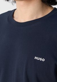 Gros plan d'une personne portant un T-shirt bleu marine à col rond avec "HUGO" imprimé en blanc sur le côté gauche de la poitrine.