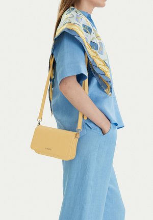 Mujer con conjunto azul claro y bufanda estampada sobre los hombros, llevando un pequeño bolso amarillo cruzado, con la mano en el bolsillo, vista lateral.