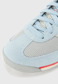 Világoskék velúr sportcipő szürke hálós panelekkel, illeszkedő fűzőkkel és vörös csíkkal a talp közelében, tapintott gumitalppal.