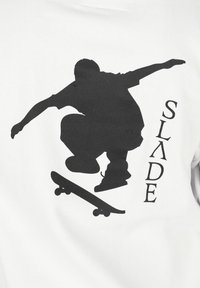 Maglietta bianca con una grande silhouette nera di uno skateboarder in pieno salto, con la parola "SLADE" stampata accanto al grafico.
