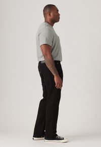 Grijze T-shirt met korte mouwen, zwarte jeans en zwarte sneakers. Zichtbare tatoeage op de arm. Zijaanzicht toont een relaxte pasvorm en eenvoudig ontwerp.