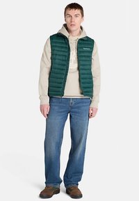 Groen donzen vest over een beige hoodie, gecombineerd met blauwe jeans met rechte pijpen en bruine schoenen. Gewatteerd ontwerp met ritssluiting en logodetail.