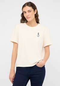 Koszulka z krótkim rękawem w kolorze kremowym, wykonana z bawełny, z małym granatowym logo na klatce piersiowej i luźnym krojem. Połączona z ciemnoniebieskimi dżinsami.