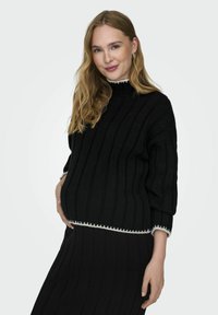 Pull en tricot noir avec une texture côtelée et un col montant, présentant une bordure claire à l'ourlet. Associé à une jupe noire assortie.