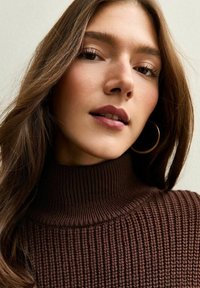 Pull à col roulé marron avec une haute col et un tricot texturé. Présente de longs cheveux doux et une boucle d'oreille en forme de cercle en or.