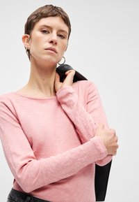 Svetloružový top s dlhým rukávom z mäkkej tkaniny s širokým výstrihom. Model drží čiernu koženú bundu cez rameno, ktorá ukazuje textúrované detaily.