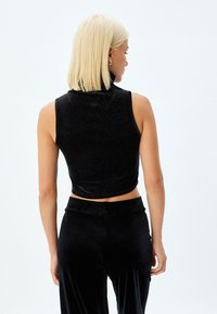 Top corto aderente senza maniche di colore nero con colletto alto, realizzato in un tessuto testurizzato e luccicante. Indossato con pantaloni neri abbinati.
