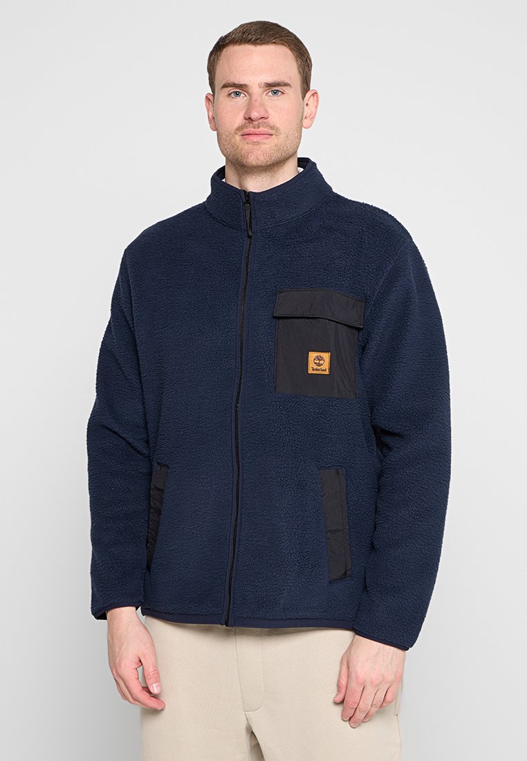 Giacca in fleece blu navy con colletto alto, chiusura con zip, accenti in nylon nero e tasca a toppa sul petto. Texture morbida.