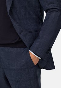 Jūras spēku krāsas šaha uzvalks ar teksturētu audumu; tam ir slaids piegriezums, šķēlumi un viens pogas aizvērums. Iekļauts balts krekla aproces akcenta elements.