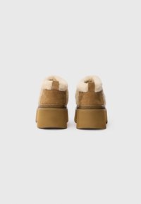 Botas de tobillo de ante marrón con forro de piel de oveja crema y una suela de goma gruesa color marrón. Presentan una pequeña lengüeta en la parte trasera y un detalle texturizado.
