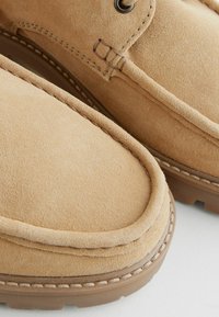 Mocasines de gamuza color arena con punta redonda, que presentan detalles de costura y una suela de goma. La textura parece suave y ligeramente aterciopelada.
