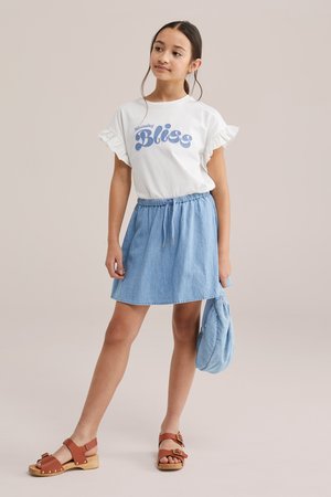 Noor tüdruk seisab, seljas valge paeltega varrukatega T-särk, millel on tekst "Bliss", sinine teksaseelik, pruunid sandaalid ja käes helesinine kott.