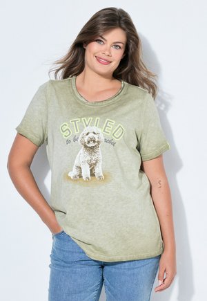 Donna con i capelli lunghi che sorride, indossa una maglietta verde con l'immagine di un cane e la scritta "STYLED to be a rebel", abbinata a jeans blu.