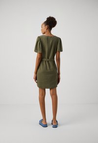 Vestido de linho verde-oliva com mangas curtas bufantes, cintura ajustada e barra até ao joelho. Combinado com sapatos azuis de slip-on.