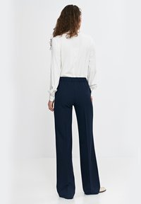 Biała bluzka z długim rękawem i elastycznym pasem w połączeniu z granatowymi spodniami o szerokich nogawkach. Gładki materiał, luźny krój i subtelne guziki na rękawach.