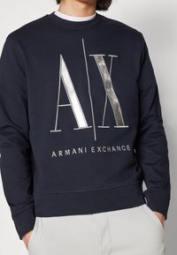 Felpa blu navy con collo a giro, con logo "AX" in argento metallico e testo "ARMANI EXCHANGE" sul davanti. Tessuto morbido.