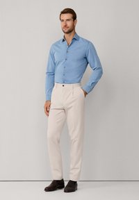 Muškarac stoji uz običan zid, odjeven u svijetloplavu košulju dugih rukava sa dugmadi, bež hlače i tamnosmeđe cipele.