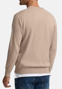 Suéter de punto beige con cuello redondo, mangas largas y puños acanalados. Textura suave con un corte ajustado, superpuesto a una camisa blanca.