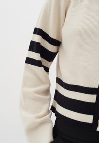 Beige ribbad cardigan med svarta horisontella ränder. Har en kort design och knäppning, vilket visar upp stickad textur.