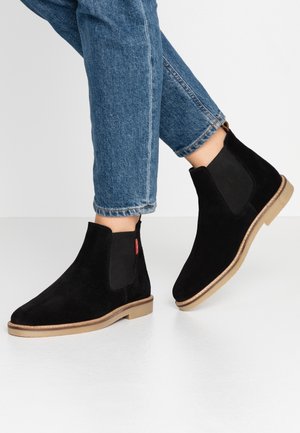 Person iført sorte Chelsea-støvler i ruskind med brune såler og opsmøgede blå jeans mod en enkel hvid baggrund.