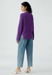 Lila gestrickter Pullover mit Raglanärmeln, seitlichen Schlitzen und einem texturierten Muster, kombiniert mit hellblauen Jeans und cremefarbenen Slingback-Heels.