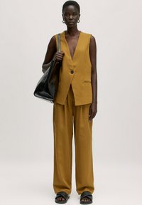 Mosterdgele mouwloze blazer en wijde broek, met een gestructureerde stof, gecombineerd met een zwarte glanzende totebag en zwarte sandalen.