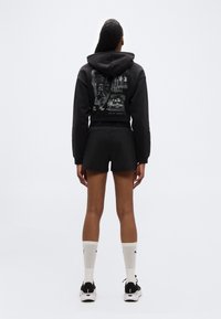 Γυναίκα που φοράει μαύρο κοντό φούτερ με στάμπα, μαύρο σορτς, άσπρες κάλτσες και μαύρα αθλητικά παπούτσια, στέκεται πλάτη σε απλό φόντο.