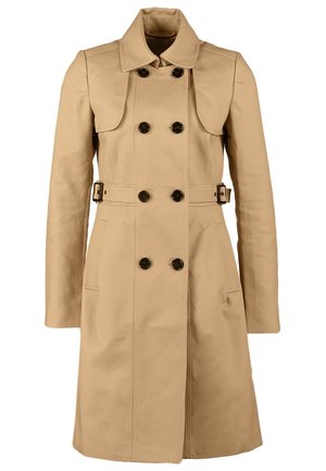 Woman wearing beige double-breasted trench coat, black pants, black ankle boots, holding a small black clutch in one hand.