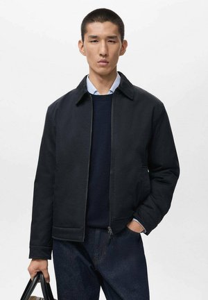Chaqueta negra con cremallera, cuello tipo solapa, bolsillos laterales y corte relajado, llevada sobre un suéter azul marino y una camisa de color azul claro, combinada con jeans oscuros.