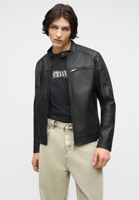 Young person with shoulder-length hair wearing a black leather jacket over a black Nirvana t-shirt and light beige jeans.