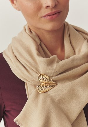 Vrouw draagt een beige sjaal vastgezet met een gestructureerde gouden broche in de vorm van een hart, over een donker bordeauxrode top.