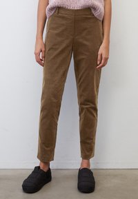 Vrouw in een bruine corduroy broek, zwarte instapquilt schoenen en een lichtpaarse gebreide mouwloze trui, staande tegen een witte muur.