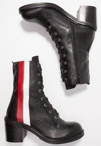 Bottes noires en cuir jusqu'à la cheville avec des talons blocs, des lacets à l'avant, des fermetures éclair sur le côté, et des rayures verticales rouges et blanches sur le côté extérieur de l'une des bottes.