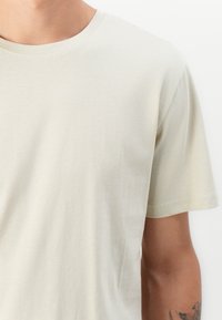 Camiseta de algodón color beige claro con escote redondo y mangas cortas. Textura suave y costuras mínimas, sin patrones o acentos visibles.