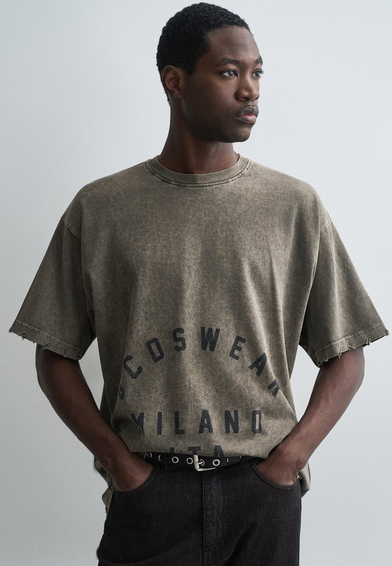 Tricou oversized de culoare maro cu margini deteriorate și logo imprimat "CDSWEAR MILANO ITA". Purtați cu blugi negri și o curea neagră cu accente din metal.