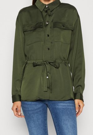 Femme portant une chemise boutonnée vert olive avec des poches poitrine et une ceinture à la taille, associée à un jean bleu.