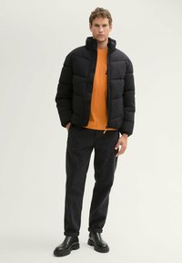 Sort puffergård med høj krave, lynlås lukning og quiltet tekstur, iført over en orange skjorte og parret med mørke jeans og sorte støvler.