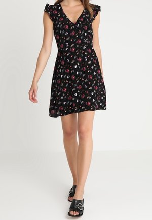 Frau geht in einem schwarzen, kurzen Blumenkleid mit flatternden Ärmeln und schwarzen offenen Sandalen mit Schnallendetail.