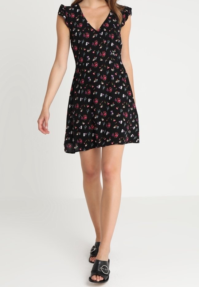 Femme marchant dans une robe noire courte à fleurs avec des manches volantées et des sandales noires à bout ouvert avec un détail de boucle.