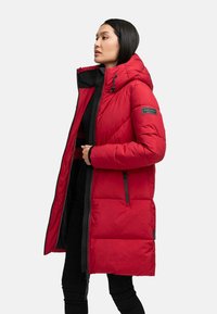 Woman wearing a long red padded coat with a hood, zipper open, standing in profile against a plain background.