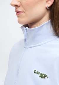 Femme portant un haut zippé bleu clair avec un col montant et un logo Lacoste vert sur la poitrine, visible de profil.
