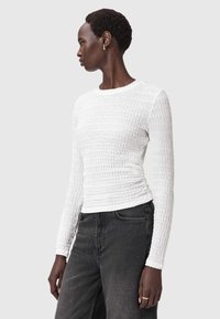 Witte lange mouwtop met een gestructureerd, gerimpeld ontwerp, ronde halslijn en getailleerde pasvorm, gecombineerd met donkere jeans voor contrast.