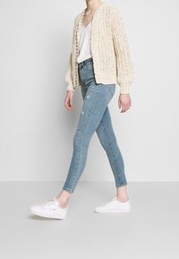 Beige gebreide cardigan met ballonmouwen, gedragen over een witte top, gecombineerd met lichte blauwe skinny jeans en witte sneakers.