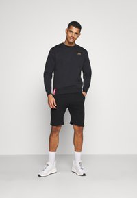 Black sweatshirt with a gold logo, paired with black shorts. Worn with white sneakers and white socks. Clean, minimalist design.