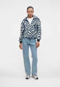 Pulover din fleece cu un model floral alb și bleumarin, cu fermoar pe jumătate în față și manșete elastice. Asociat cu blugi în dungi albastre deschise și adidași.