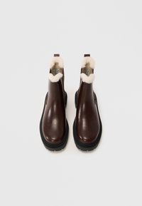 Hnědé kožené kotníkové boty s tlustou černou podrážkou a bílým fleece podšívkou viditelnou na horních okrajích, zobrazené na bílém pozadí.