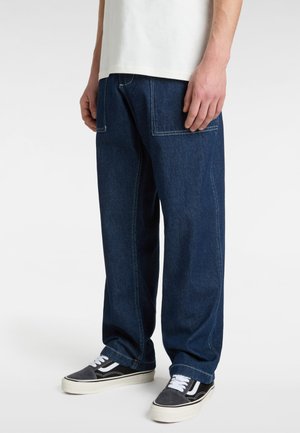 Dunkelblaue, weit geschnittene Jeanshosen mit sichtbaren Nähten, zwei Vordertaschen und lässigen schwarz-weißen Sneakers. Das Material wirkt robust und strukturiert.
